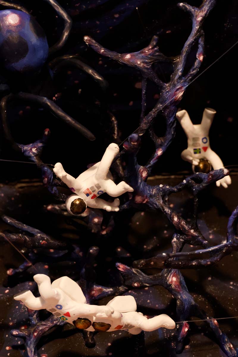 A slightly different angle of the astronaut who sits near the spider. In the background, an upside-down astronaut flails their arms. The entwined duo floats near the bottom of the web.