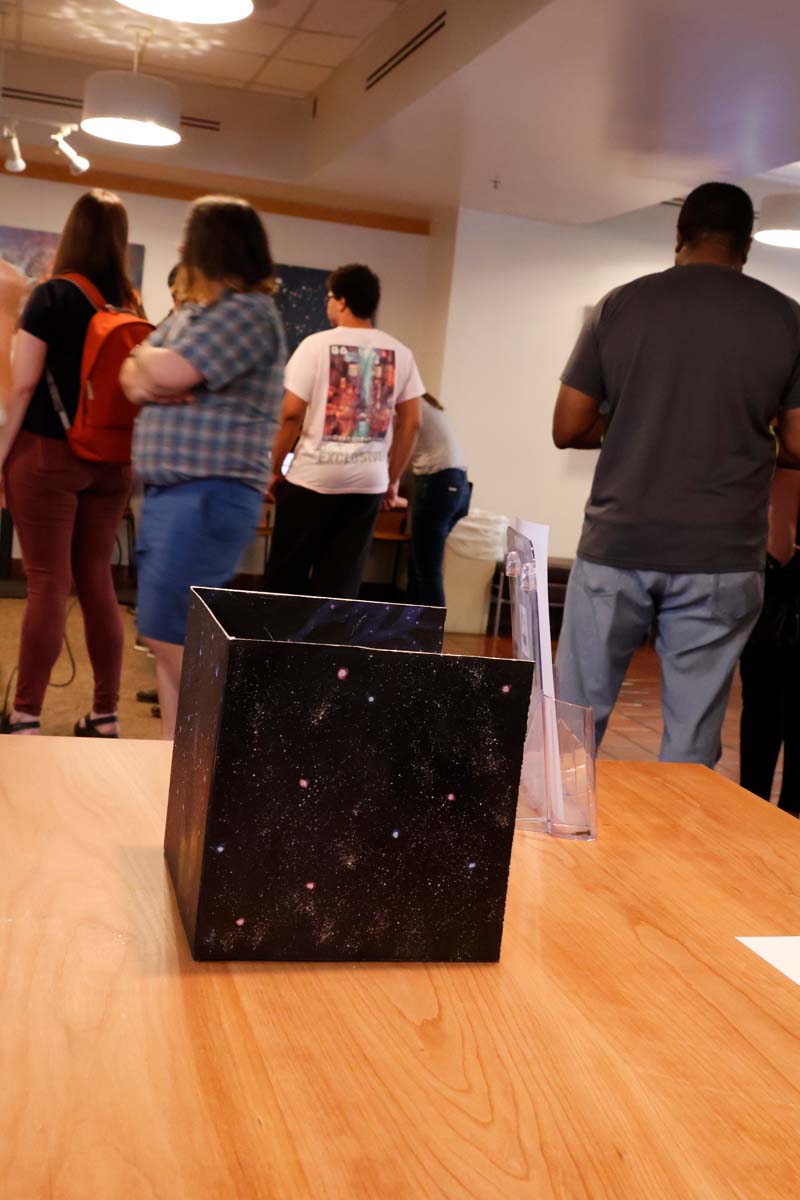 The cosmic web box sitting on a table. From this angle, two exterior faces and the open top of the box are visible. The pattern on the outside is slightly different; the outside shows clusters of stars, while the inside adds additional brush strokes to connect these into filaments.