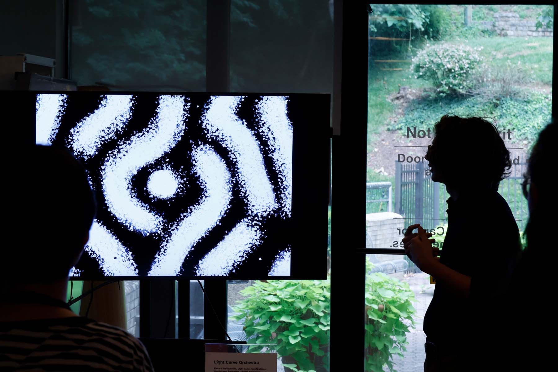 A silohetted student describes his animation to a group of onlookers, the animation is playing on a large television screen.
