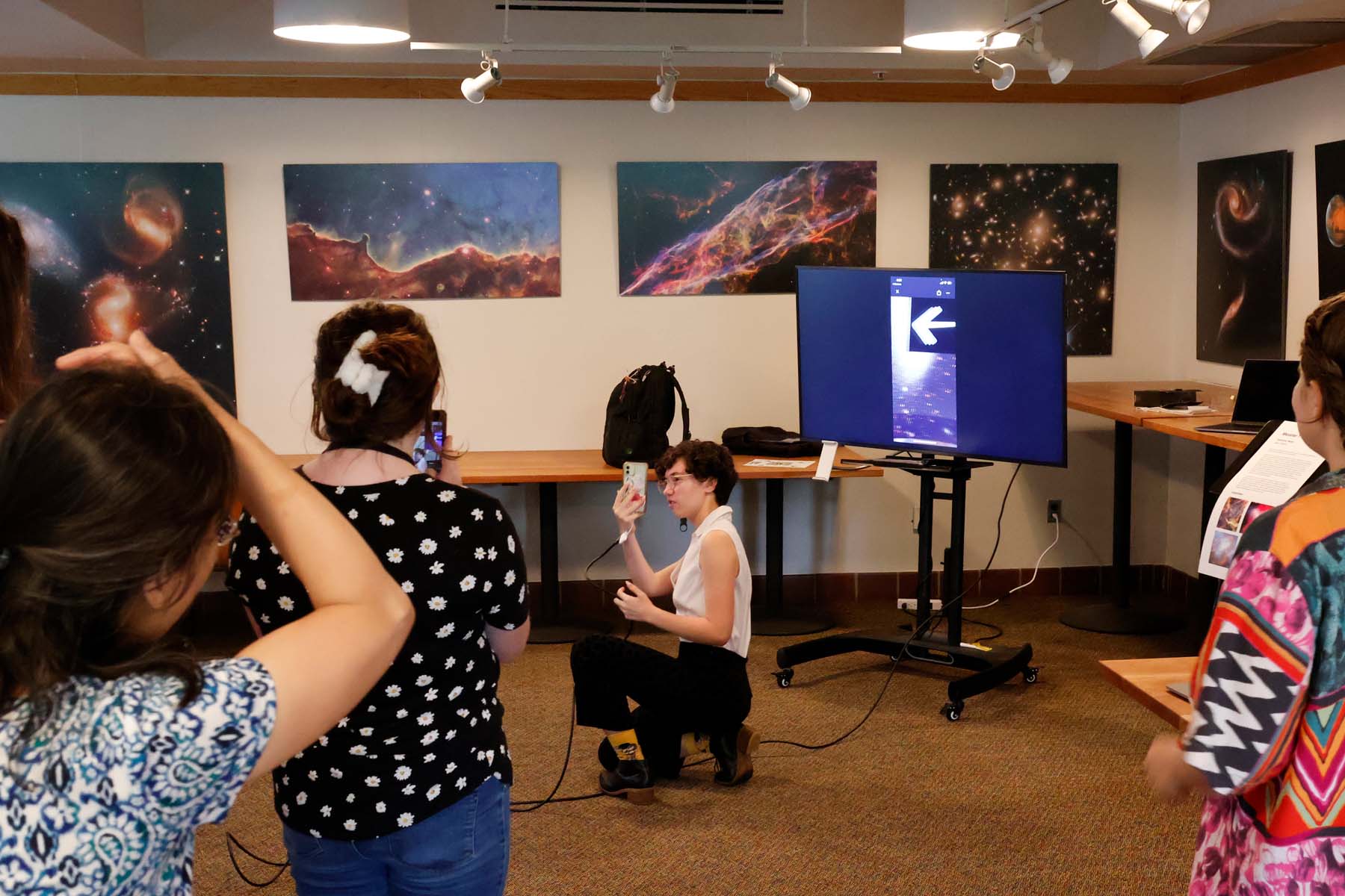 A student kneels looking at a smartphone, what is on her screen is displayed on the television behind her. A group of people watch, and one takes a video with her phone.