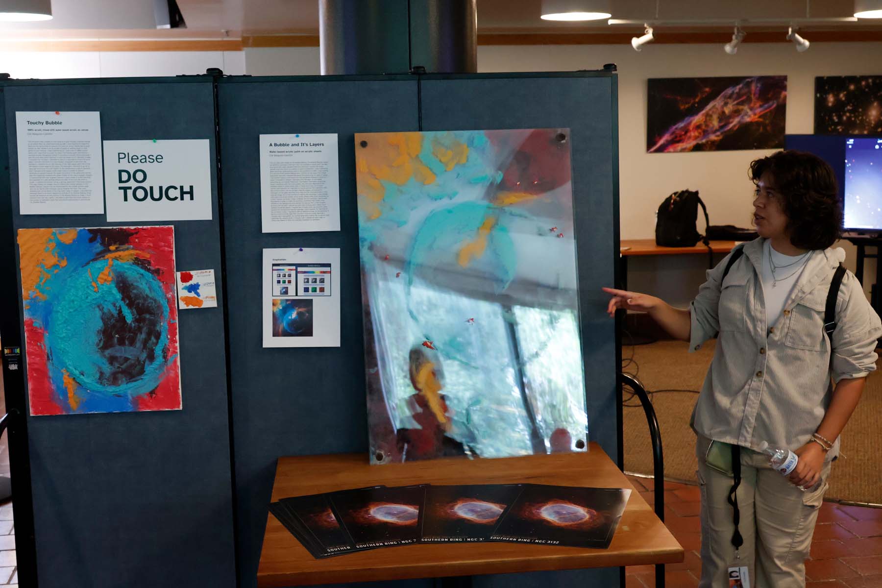 A student points towards a piece that shows the bubble nebula; the piece is layered plexiglass that has been painted and stacked to give it a textured look. To the left is a more traditional "oil on canvas" version of the piece