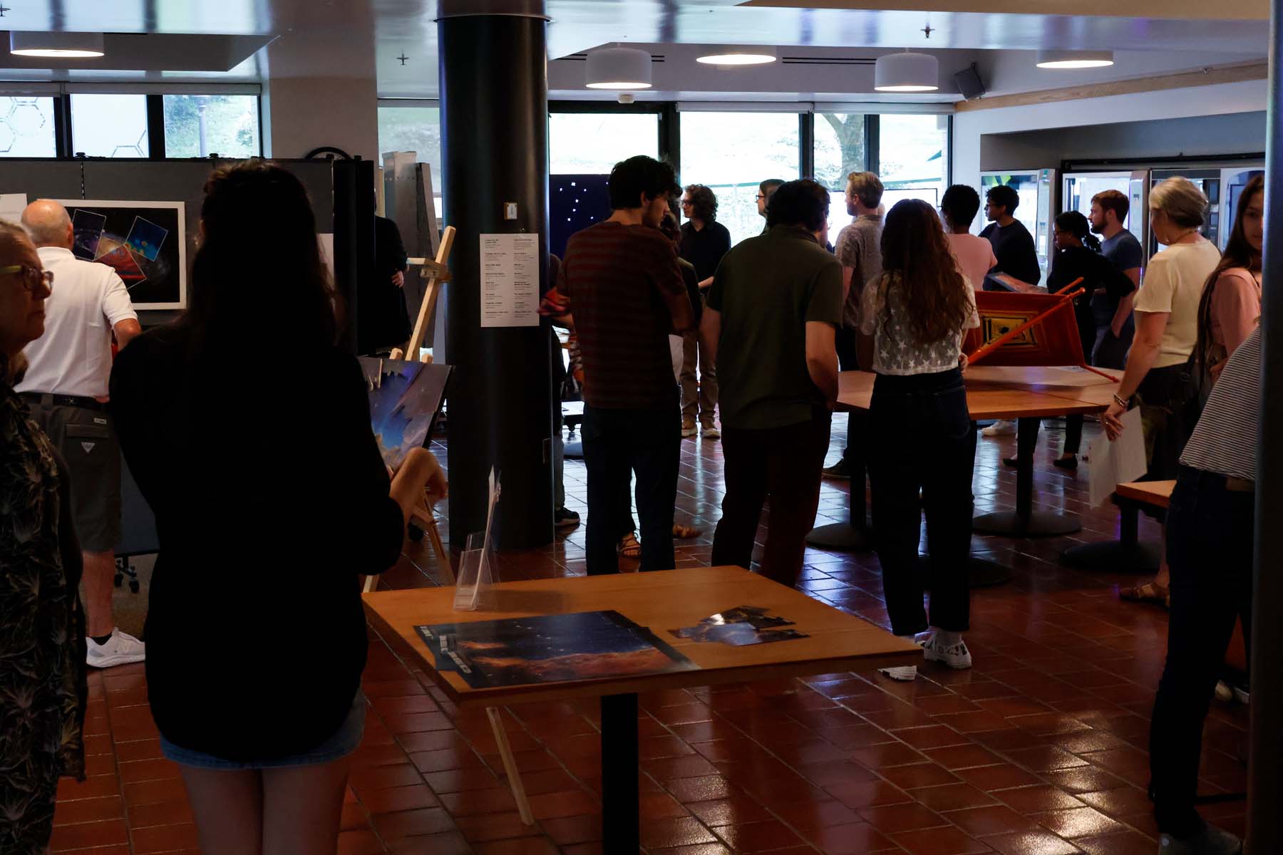 Approximately two dozen people gather around a menagerie of pieces. A centrally located table contains JWST release image posters.