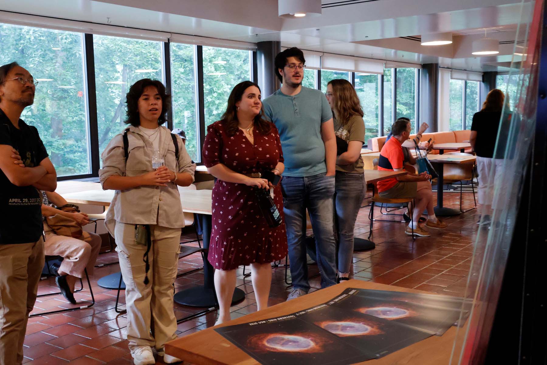 Photograph of several people reading the description of an art piece. Behind them is a row of tables and wall of windows on a forest. In front of them, several posters of space imagery sit on a table.